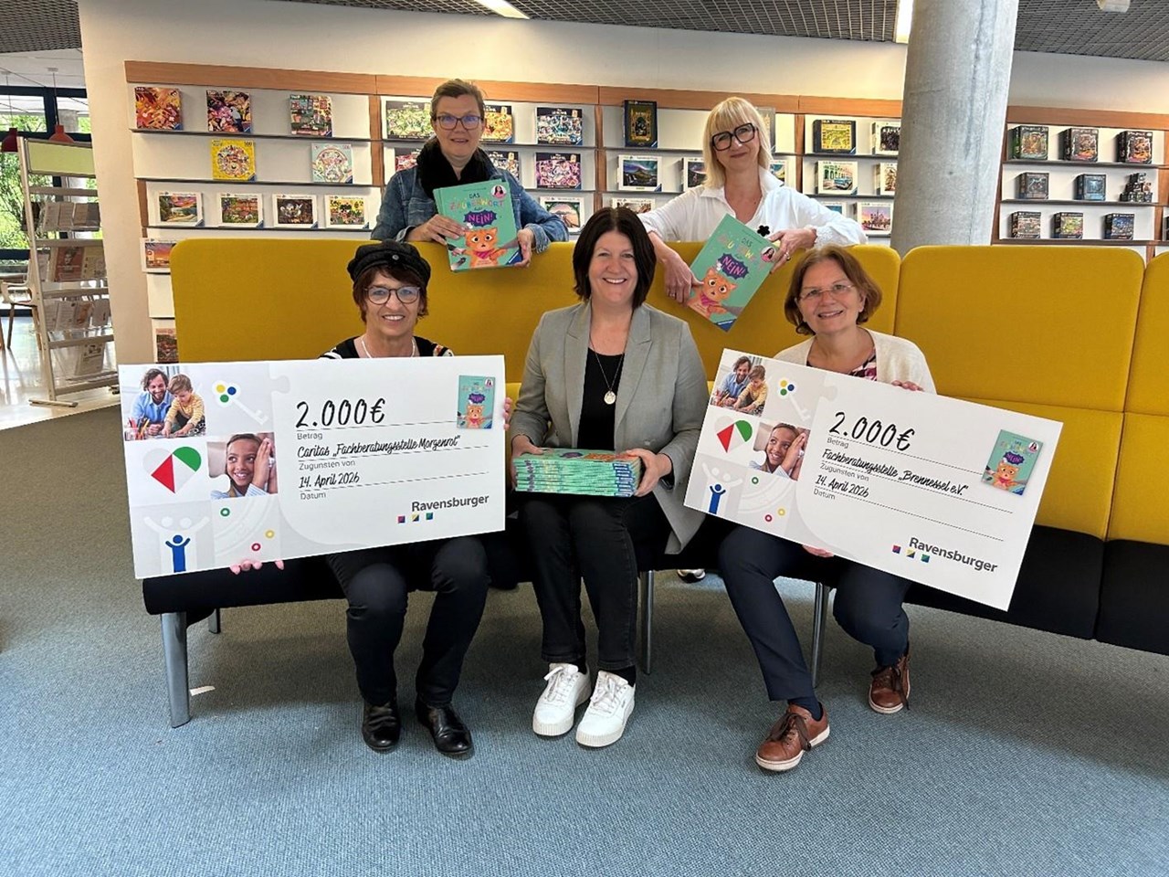 5 Frauen haben sich um ein großes Lesesofa positioniert und präsentieren zwei große Spendenschecks - im Hintergrund ein Bücherregal mit Ravensburger Produkten.